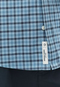 Blå og sort ternet skjortestof med en synlig label, der læser "Durable Goods Lot 082" nær kanten, knappet ved kraven.