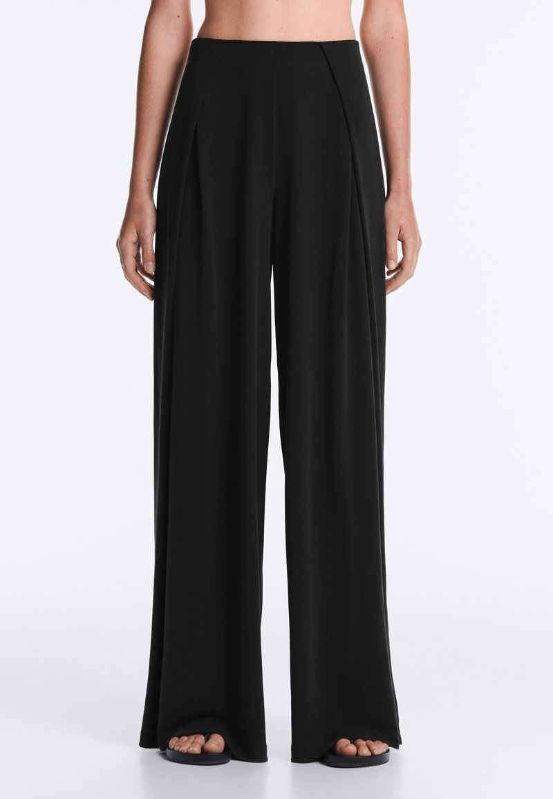 Femme portant un pantalon noir taille haute à jambes larges associé à des sandales noires, debout devant un fond clair uni.
