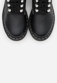 Botas de cuero negro hasta el tobillo con puntas redondas, ojales plateados y costura blanca en contraste a lo largo de la suela. Los cordones son negros y texturizados.