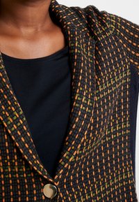 Blazer noir texturé avec un motif à carreaux comportant des accents orange et vert, et un bouton en argent. Top noir visible en dessous.