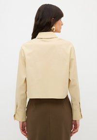 Camicia beige tagliata, con colletto classico, polsini attillati e texture liscia, abbinata a una gonna marrone scuro.