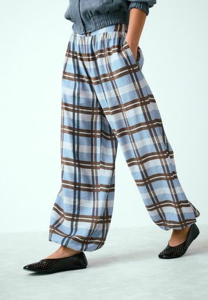 Person wearing loose blue and brown plaid pants with hands in pockets and black perforated flats, standing on light floor.