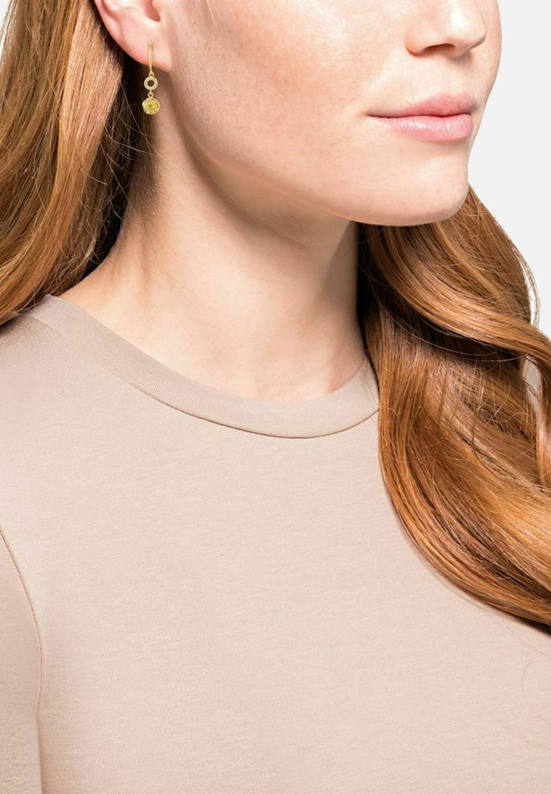 Guldhängande örhängen med cirkulär design, med en strukturerad yta och en slät finish, bärs med en ljusbeige topp.