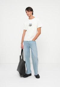 Biała bawełniana koszulka z graficznym nadrukiem, zestawiona z jasnoniebieskimi dżinsami o prostym kroju. Modelka trzyma czarną skórzaną torbę typu tote. Czarne buty.