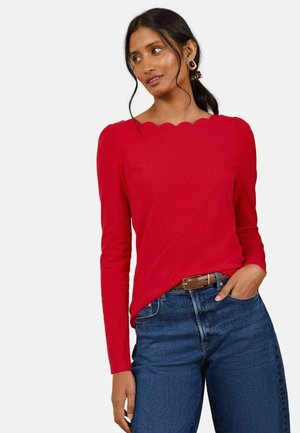 Kvinde med mørkt hår iført en rød sweater med bølget halsudskæring, blå jeans med brunt bælte og store firkantede øreringe, hånd i lommen.