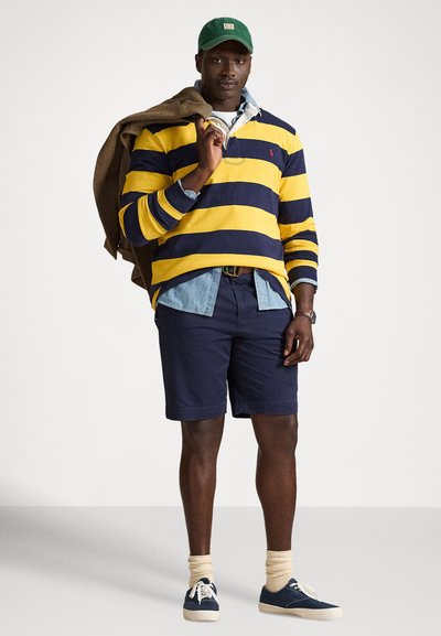 Zöld sapkát viselő férfi, sárga és tengerészkék csíkos rögbi ingben farmering felett, tengerészkék rövidnadrágban, krémszínű zokniban, kék tornacipőben, barna dzsekit vállára dobva.