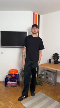 Μαύρο κοντομάνικο μπλουζάκι, μαύρο αθλητικό παντελόνι και μαύρα αθλητικά παπούτσια με λευκές ρίγες. Χαλαρή εμφάνιση με τατουάζ στα χέρια. Πολύχρωμο φωτιστικό κοντά.