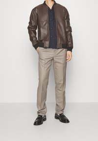 Giacca bomber in pelle marrone con polsini a costine neri, abbinata a pantaloni eleganti beige e scarpe in pelle nere. La giacca presenta una chiusura con zip.