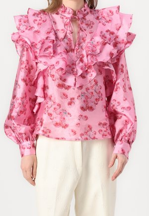 Różowa kwiatowa bluzka z dużymi falbanami na ramionach i klatce piersiowej, długimi bufkami na rękawach oraz wiązaniem pod kołnierzykiem, w zestawieniu z białymi spodniami o fakturze.