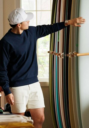 Person in navy sweatshirt and white shorts points to a row of surfboards in various colors, displayed against a bright window backdrop.