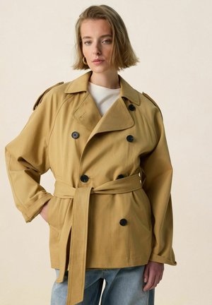 Frau mit kurzem blondem Haar, die goldene Creolen und einen beigen, doppelt geknöpften Trenchcoat mit Gürtel über hellblauen Jeans trägt.