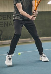 Fekete sportnadrág és fehér sportcipő fekete és sárga akcentusokkal, egy teniszütőt tart a kezében felkészült állásban egy kék teniszpályán.