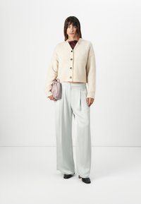 Lys beige cropped cardigan med knapper, parret med grå brede bukser og en blød lavendel taske. Modellen bærer sorte hæle.