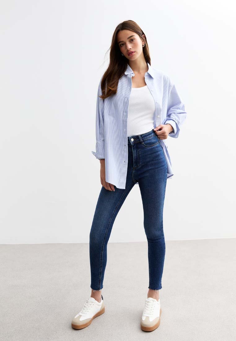Camisa de botón azul y blanca a rayas clásica, top blanco ajustado, jeans skinny azul oscuro y zapatillas blancas con suelas marrones.