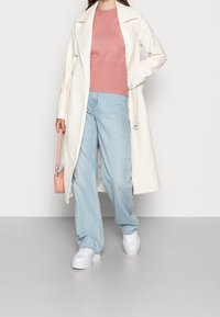 Trench-coat blanc, pull côtelé rose, jean large bleu clair, et baskets à plateforme blanches. Un petit sac à main rose complète la tenue.