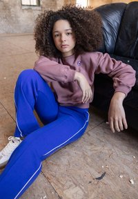 Jongere met krullend haar zittend op de vloer naast een zwart leren bank, gekleed in een paarse sweatshirt en blauwe trainingsbroek met witte sneakers.