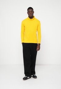 Pulover galben cu mâneci lungi și guler înalt, cu o textură moale și pantaloni negri. Prezintă un logo subtil pe guler și o croială lejeră.