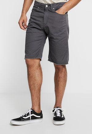 Mand iført mørkegrå knælange shorts og sort-hvide afslappede sneakers, stående med den ene hånd i lommen.