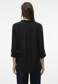 Maglietta nera a maniche lunghe con i polsini arrotolati, vestibilità rilassata, dettaglio piega sul retro e texture liscia. Tessuto leggero con un design semplice.