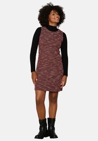 Vestido rojo texturizado con un diseño sin mangas, superpuesto a una blusa negra de mangas largas. Presenta un escote redondo y se complementa con botas negras.