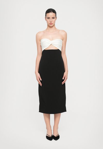 Schwarzes Midikleid mit passender Silhouette, das ein weißes, gedrehtes Bandeau-Oberteil und einen glatten, dehnbaren Stoff aufweist. Spitzige schwarze Absätze.