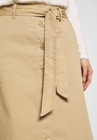 Khaki skirt with a waist tie, featuring a button front and smooth texture. Buttons are round, made of tan plastic.