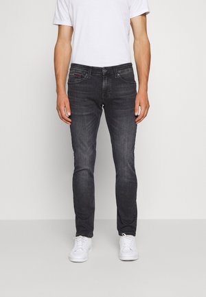 Homme portant un jean slim noir délavé, un t-shirt blanc et des baskets blanches, debout devant un fond clair uni.