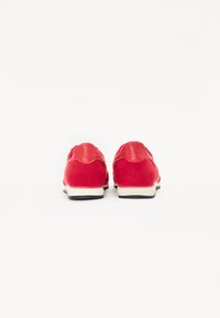 Red suede sneakers with smooth leather accents, rounded shape, contrasting white sole, and textured surface. Back view showcasing scalloped edges.
