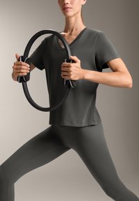 Tummanharmaa urheilupaita V-pääntiellä, yhdistettynä matching leggingseihin. Malli pitää mustaa Pilates-rengasta, jossa on kahvat.