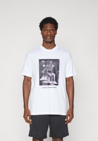 White cotton t-shirt featuring a graphic in gray tones depicting a basketball player, with “adidas basketball” text at the bottom.
