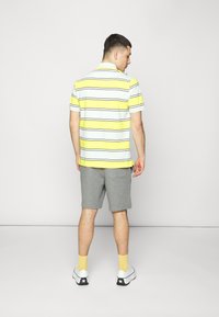 Kortermet gul og hvit stripet polo skjorte, grå shorts, hvite joggesko med svarte detaljer, gule sokker. Enkelt bakgrunn.