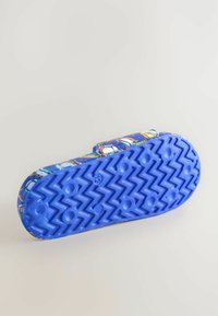 Blå gummisandal med zigzag-mönster på sulan, med en färgglad mönstrad rem och justerbar kardborrestängning. Texturerad yta för grepp.