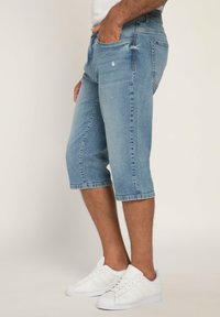 Pantalones cortos de mezclilla azul claro hasta la rodilla con un acabado desgastado, ligero desgarro y corte recto, combinados con zapatillas blancas.