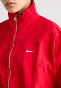 Rdeča Nike jakna z visokim ovratnikom, srebrnim zadrgo in belim logotipom na levem prsnem delu. Gladka tekstura in športen dizajn.