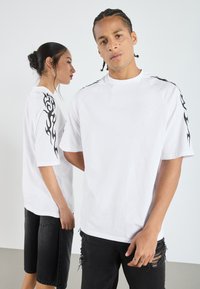 Camiseta blanca de manga corta con gráficos de llamas negras en los hombros, combinada con pantalones cortos negros desgastados. Textura suave de algodón.