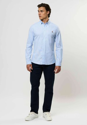 Šviesiai mėlynas marškinėliai su subtiliu languotu raštu, ilgomis rankovėmis ir mažuoju logotipu ant krūtinės, derinami su tamsiomis džinsais ir baltais sportbačiais.