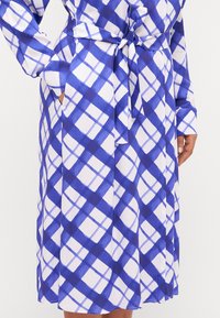 Un vestito a scacchi blu e bianco realizzato in un tessuto morbido, con maniche lunghe, una cintura in vita e tasche laterali.