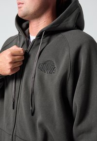 Tamnosiva majica s kapuljačom na patentni zatvarač, s teksturiranim materijalom, podesivim vezicama i izvezenim logotipom na prsima.