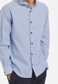 Vaaleansininen ruudullinen nappipaita, joka on valmistettu puuvillasta. Varustettu leviällä kaulalla, tummilla nappeilla, pitkillä hihoilla ja kaarevalla helmalla.