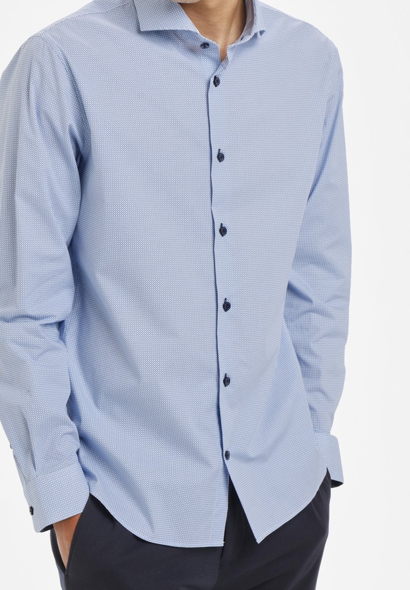 Vaaleansininen ruudullinen nappipaita, joka on valmistettu puuvillasta. Varustettu leviällä kaulalla, tummilla nappeilla, pitkillä hihoilla ja kaarevalla helmalla.