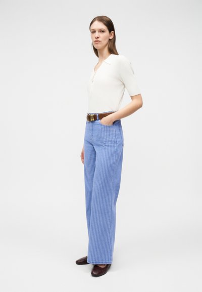 Γυναίκα που στέκεται, φορώντας λευκό πόλο με κοντά μανίκια, μπλε ριγέ παντελόνι φαρδιάς γραμμής με ψηλή μέση, καφέ ζώνη και σκούρα καφέ παπούτσια χωρίς τακούνι.