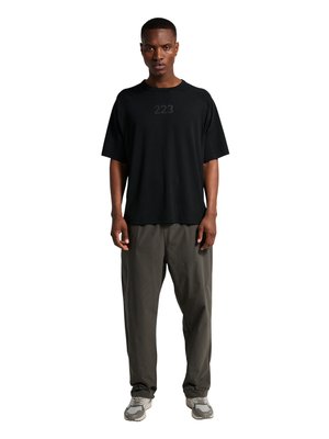 Mann, der frontal steht und ein schwarzes übergroßes T-Shirt mit der Aufschrift „223“ auf der Brust, graue lockere Hose und graue Sneaker auf weißem Hintergrund trägt.