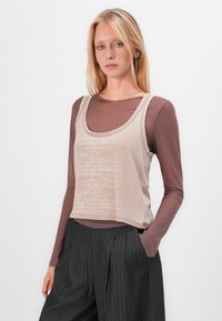 Lys beige tank topp med subtil tekstmønster, lagd over en brun langermet skjorte. Kombinert med stripete bukser, som viser en avslappet passform.