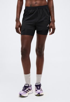 Atletisch persoon met zwarte gelaagde shorts, grijze sokken en paarse hardloopschoenen, staand tegen een effen lichte achtergrond.