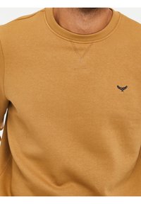 Gros plan d'une personne portant un sweat-shirt à col ras du cou jaune moutarde avec un petit logo brodé bleu marine sur la poitrine.