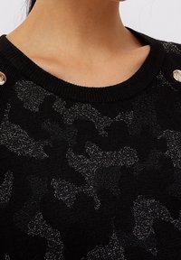 Svart stickad tröja med en texturerad, skimrande mönster, rund halsringning och metallknappar på axeln.