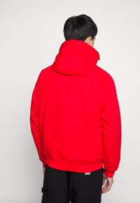 Person som står med ryggen mot betraktaren iförd en ljus röd huvenjacka och svarta byxor mot en enfärgad ljus bakgrund.