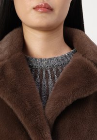 Cappotto di pelliccia marrone con un ampio colletto, abbinato a un maglione grigio lavorato a maglia con strisce verticali e un colletto a coste.