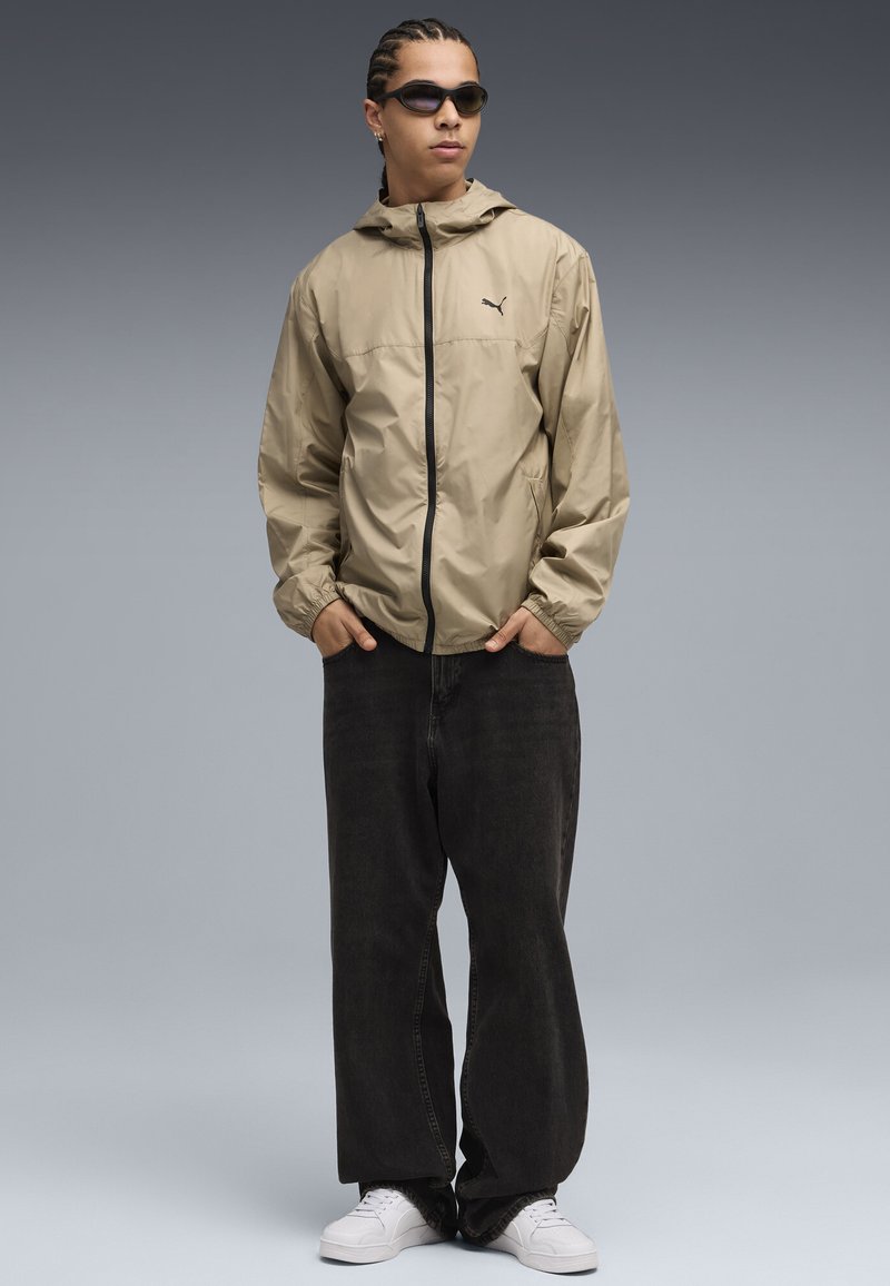 Lys beige, vandafvisende jakke med lynlås foran og hætte. Løse sorte jeans og hvide sneakers, accessoriseret med mørke solbriller.