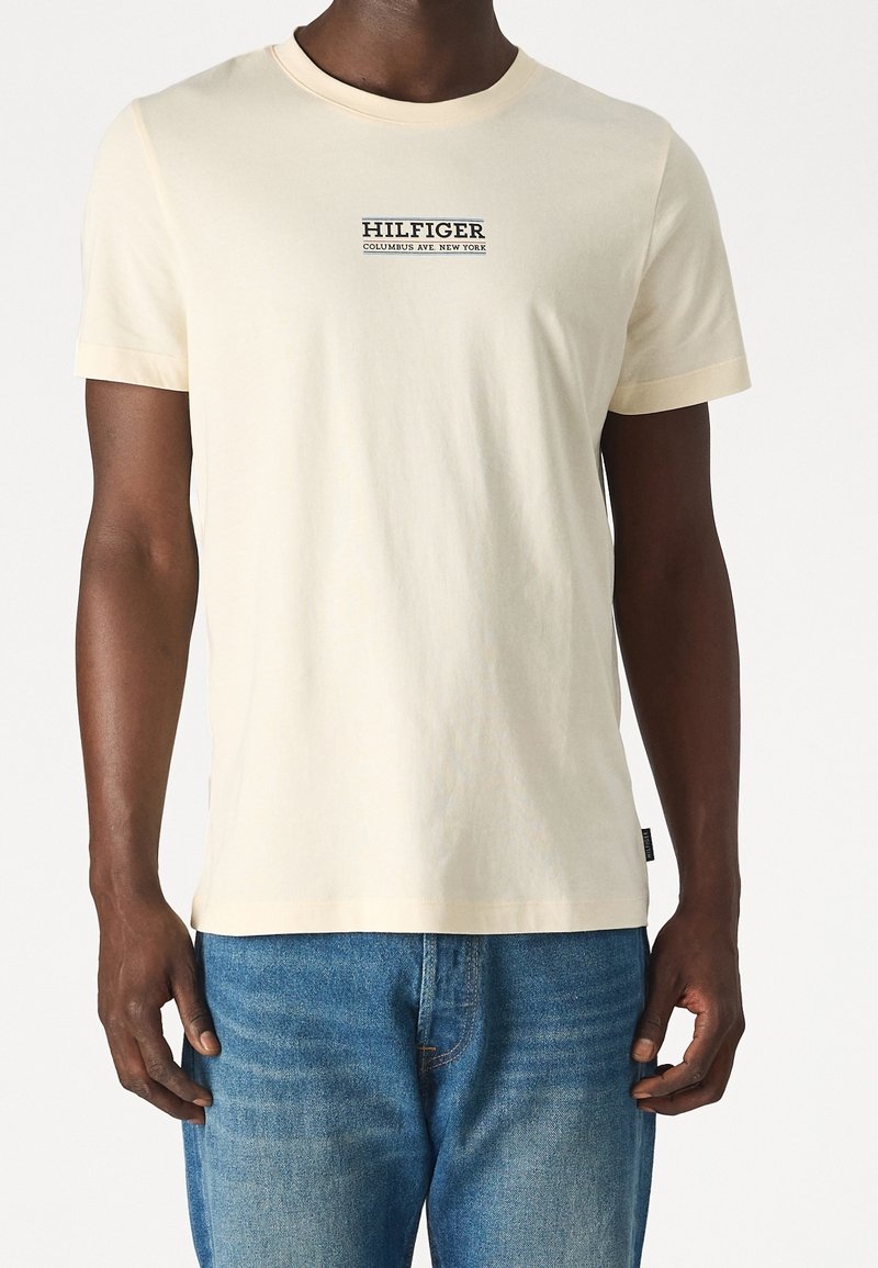 Muž v krémovém tričku s krátkým rukávem s nápisem "HILFIGER COLUMBUS AVE. NEW YORK", doplněném o modré džíny.
